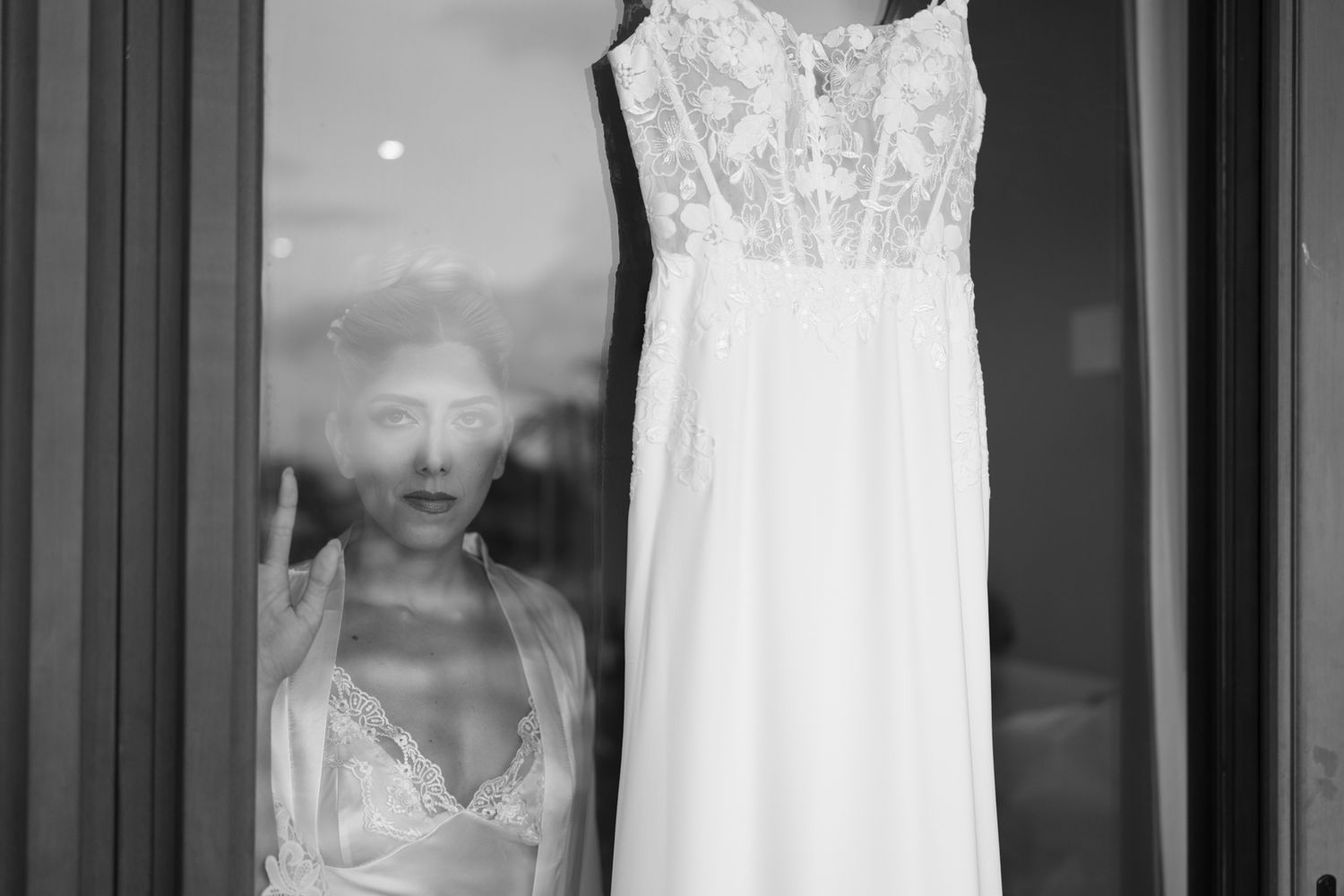 Black and white photo of a bride in lingerie looking through a window at her wedding dress hanging outside.