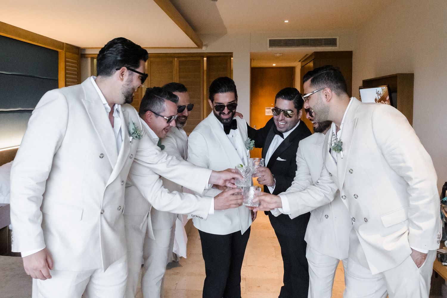 Groomsmen in white suits toasting drinks together in a hotel room before a wedding ceremony.