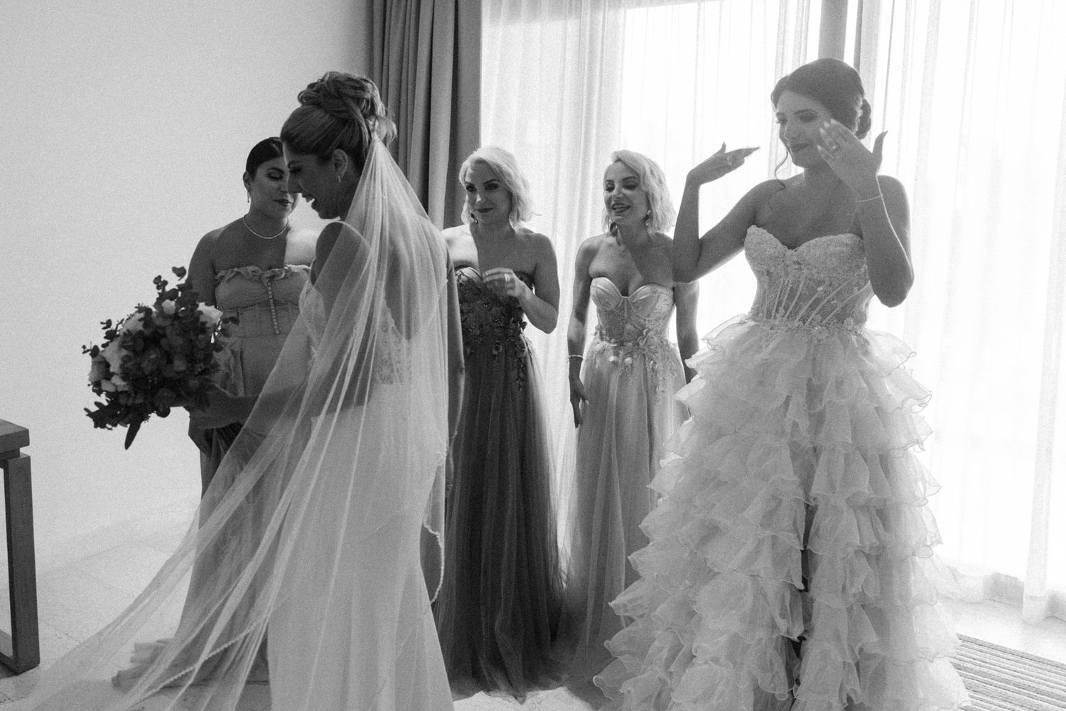 Bride and bridesmaids share emotional moment in elegant wedding gowns, black and white photo.