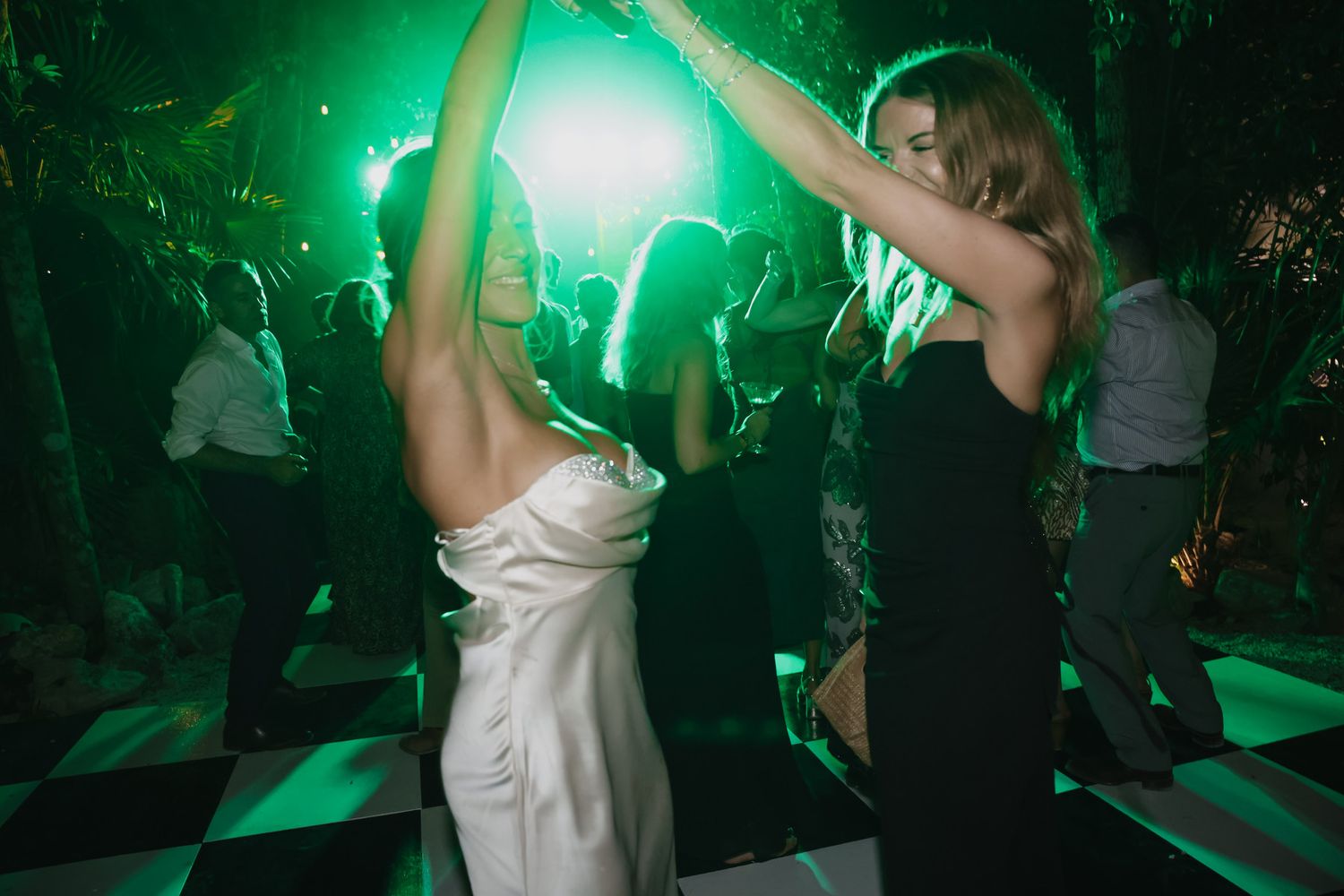 Bride in white dress dancing with guest on checkered dance floor under green lights at outdoor wedding reception.