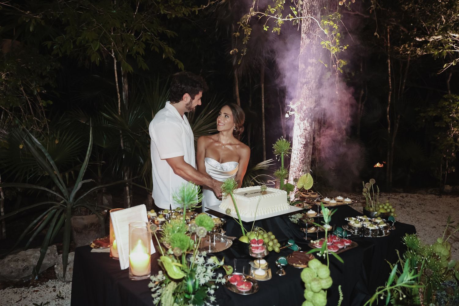Couple cutting wedding cake at night outdoor reception with purple smoke and tropical decor on black tablecloth.