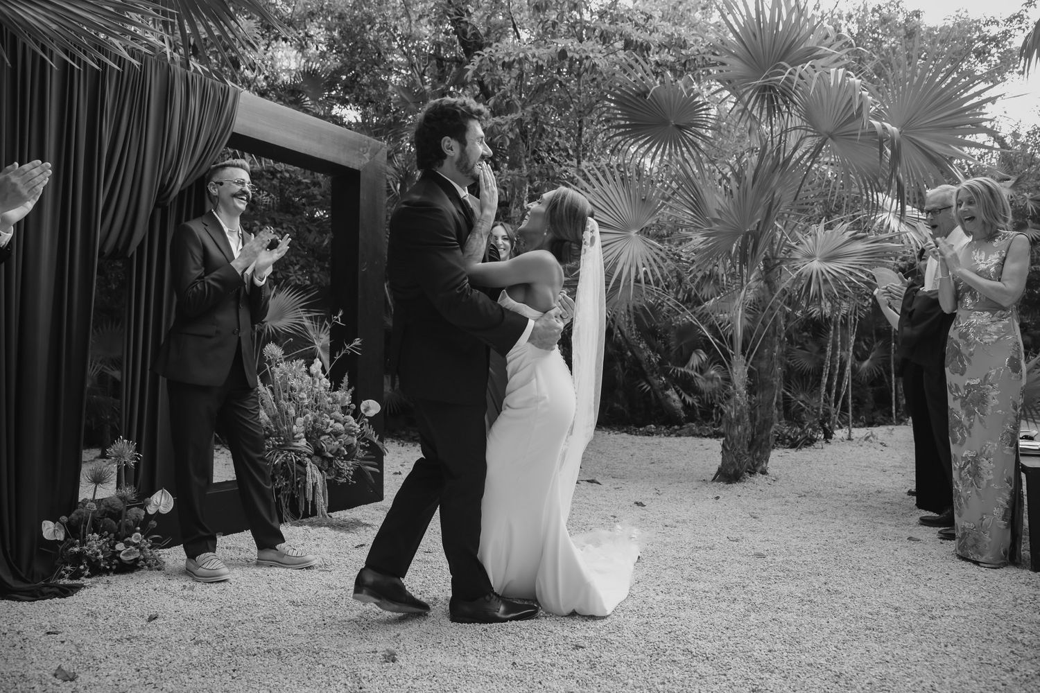 Bride and groom share first dance at outdoor wedding ceremony surrounded by tropical plants and applauding guests.