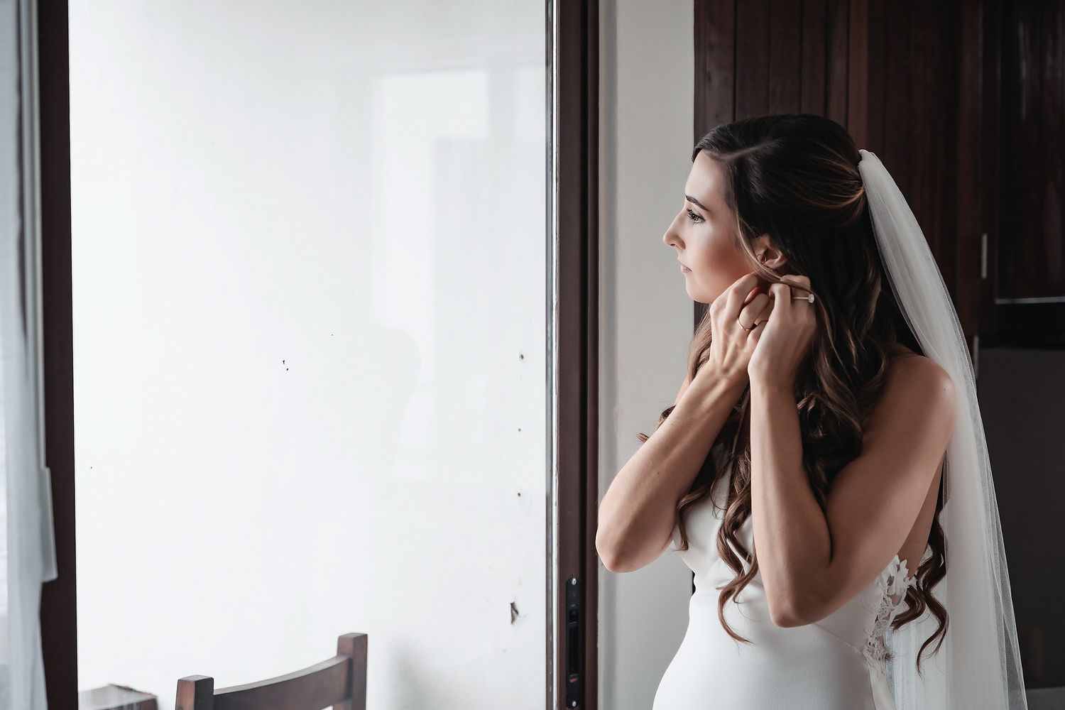 Bride in white gown and veil putting on earrings near a window on her wedding day.