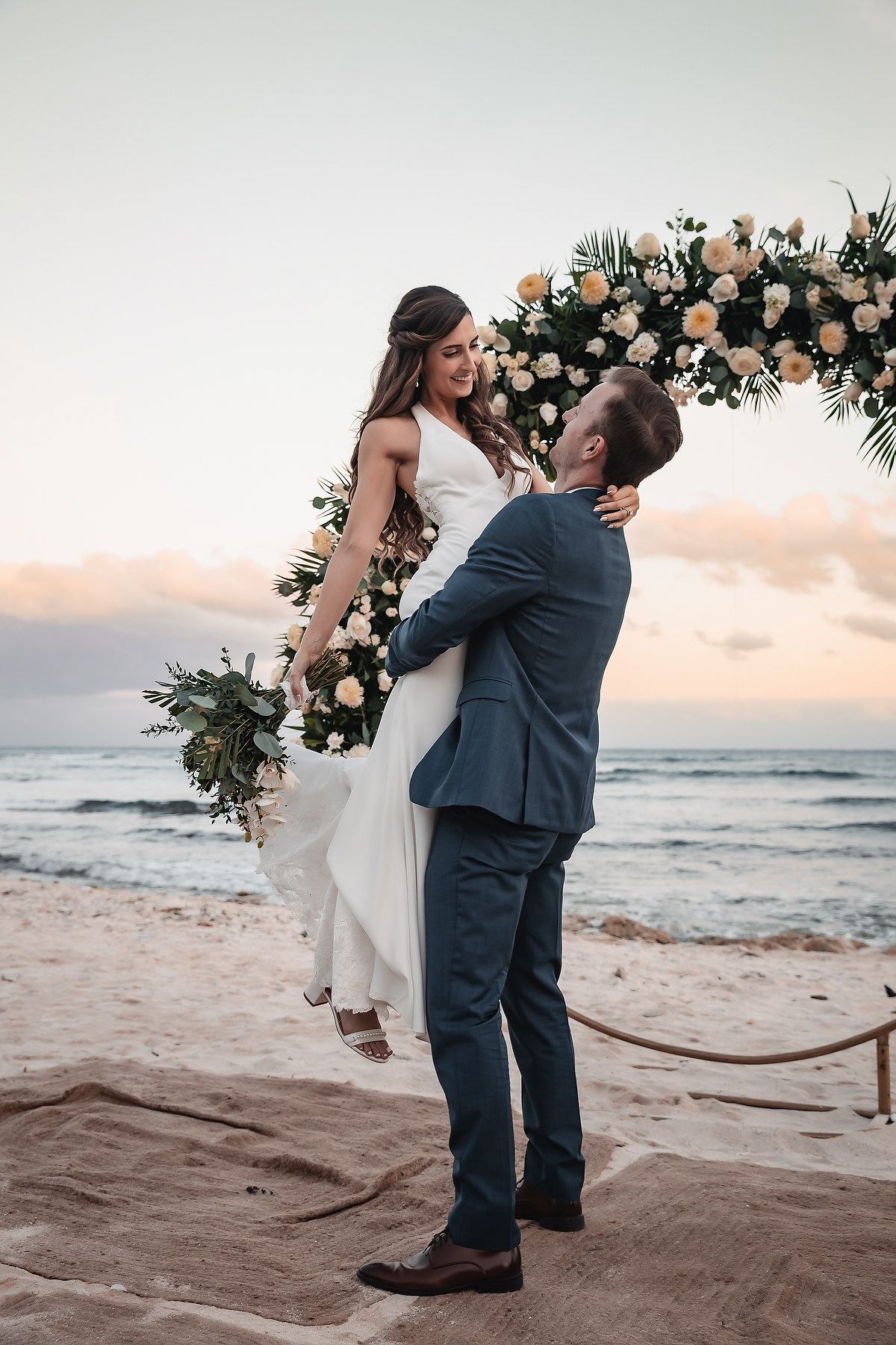 Groom lifts bride in white dress at beach wedding ceremony with floral arch at sunset.