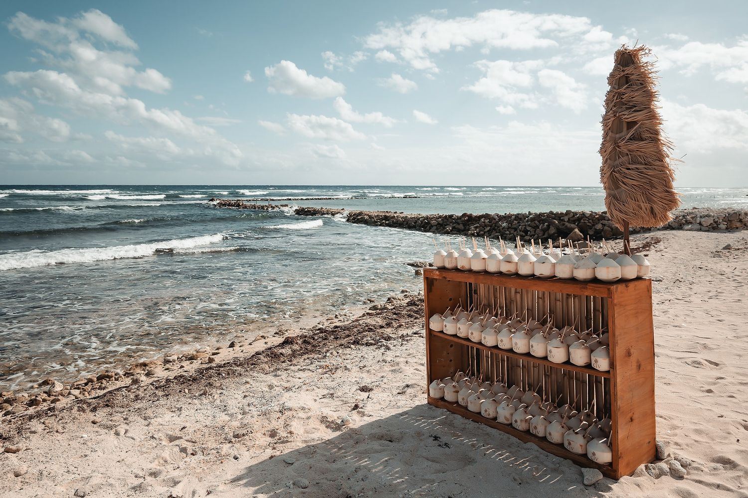 Rustic wooden bar with bottles on a sandy beach shore with ocean waves and cloudy sky backdrop.