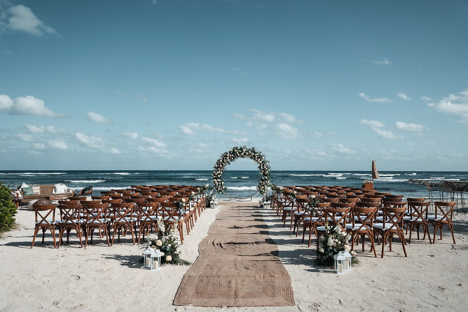 Beach wedding ceremony setup with floral arch, wooden chairs, and burlap aisle runner overlooking the ocean.