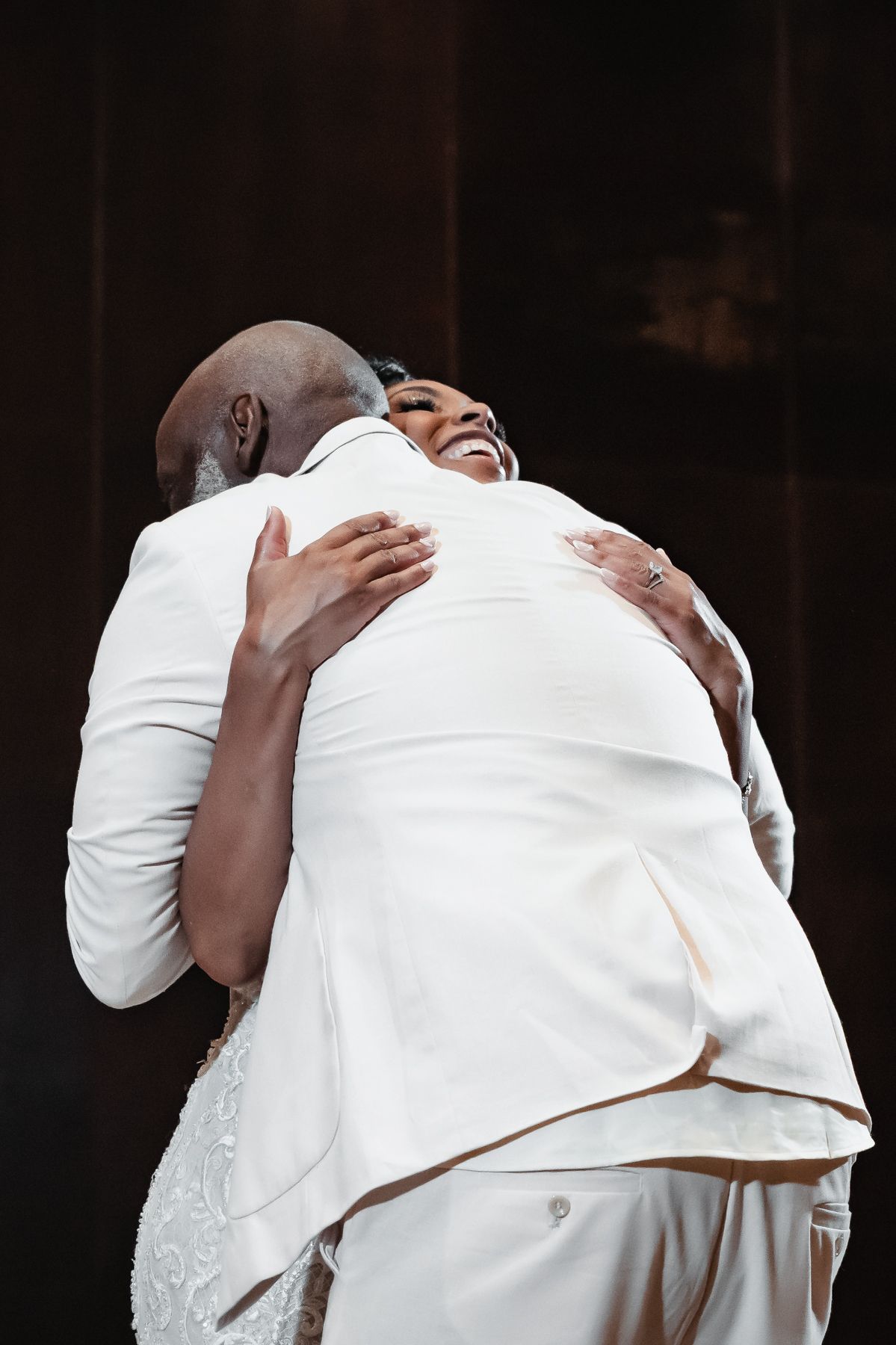 Two people in white outfits share a joyful embrace on stage, laughing and hugging warmly.