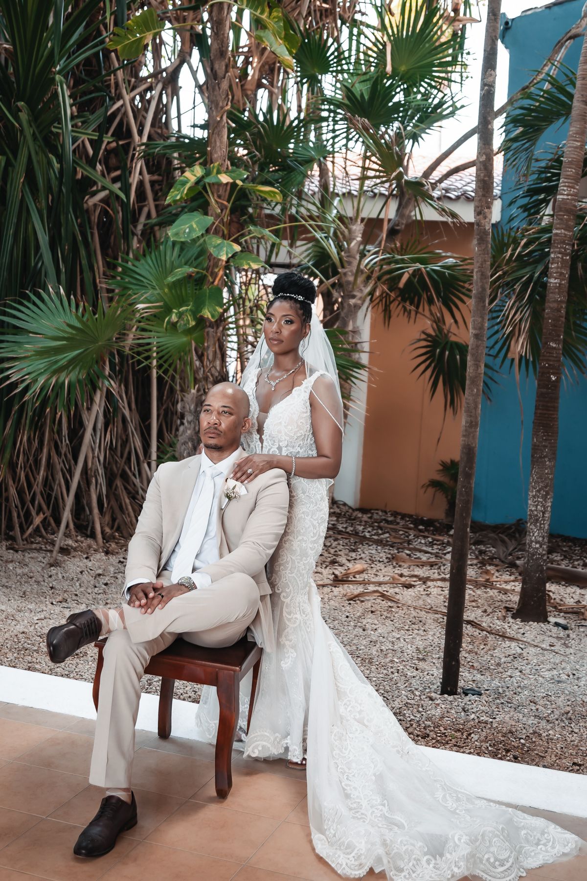 Elegant wedding couple posing outdoors, groom seated in tan suit, bride standing in lace mermaid gown with veil.