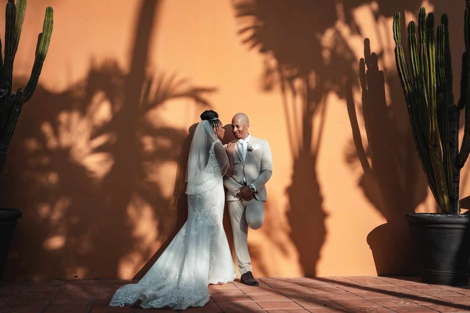 Bride and groom sharing a romantic kiss in front of a terracotta wall with palm tree shadows and cacti.