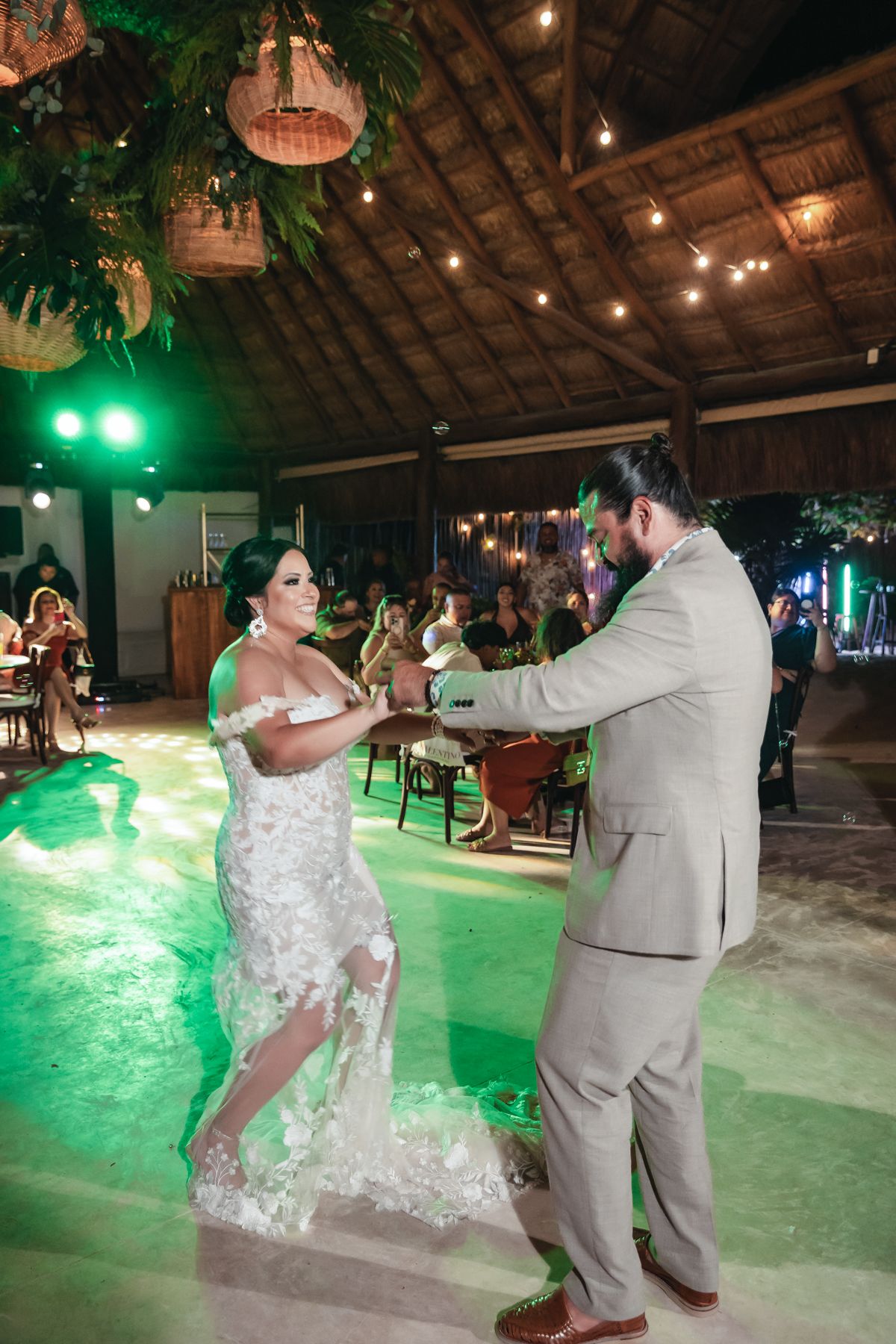 Bride and groom share their first dance under warm string lights in a rustic open-air wedding venue with green lighting.