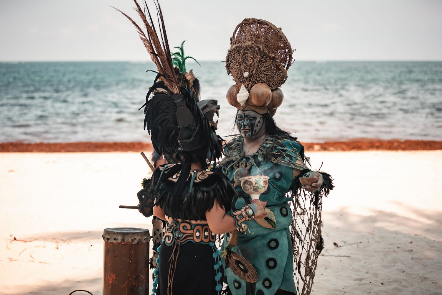 Two performers in elaborate traditional tribal costumes with feathers and masks stand on a sandy beach by the ocean.