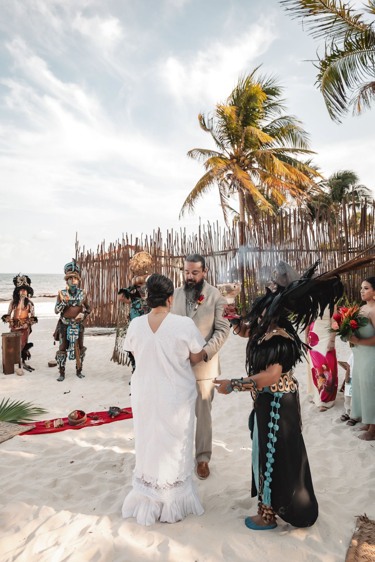 Beach wedding ceremony with couple exchanging vows, officiant in black feathered costume, palm trees, and rustic bamboo fence backdrop.
