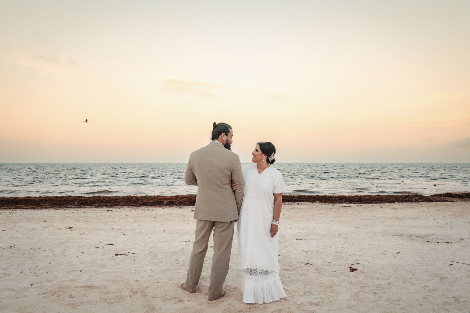 Couple in wedding attire standing on a sandy beach at sunset, facing the ocean in a romantic seaside ceremony.