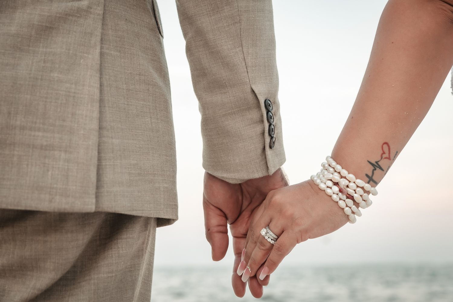 Couple holding hands at wedding, bride wearing pearl bracelet and ring, groom in beige suit, romantic outdoor ceremony.