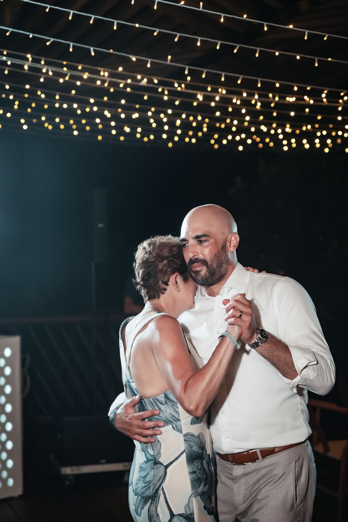 Couple shares intimate dance under warm string lights at outdoor evening wedding reception.