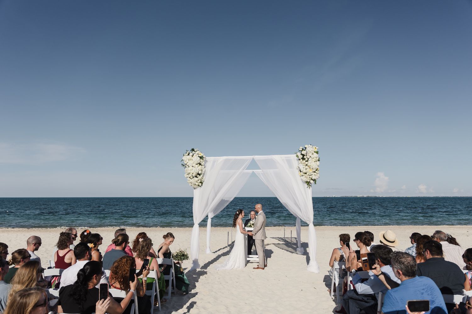 Romantic beach wedding ceremony with white floral arch, couple exchanging vows, guests seated on white sand by turquoise ocean.