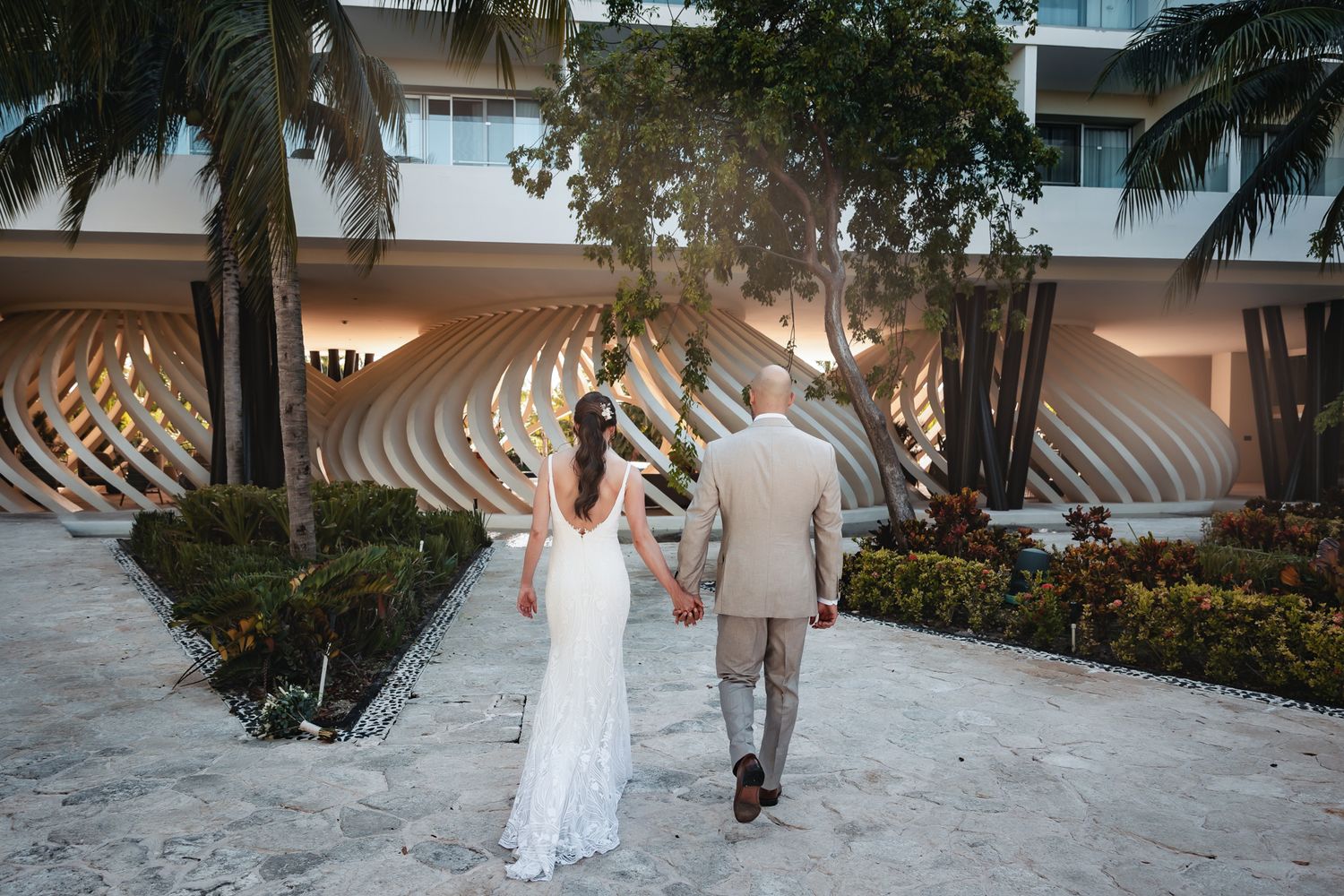 Bride and groom walking hand in hand toward a modern resort building surrounded by palm trees on their wedding day.