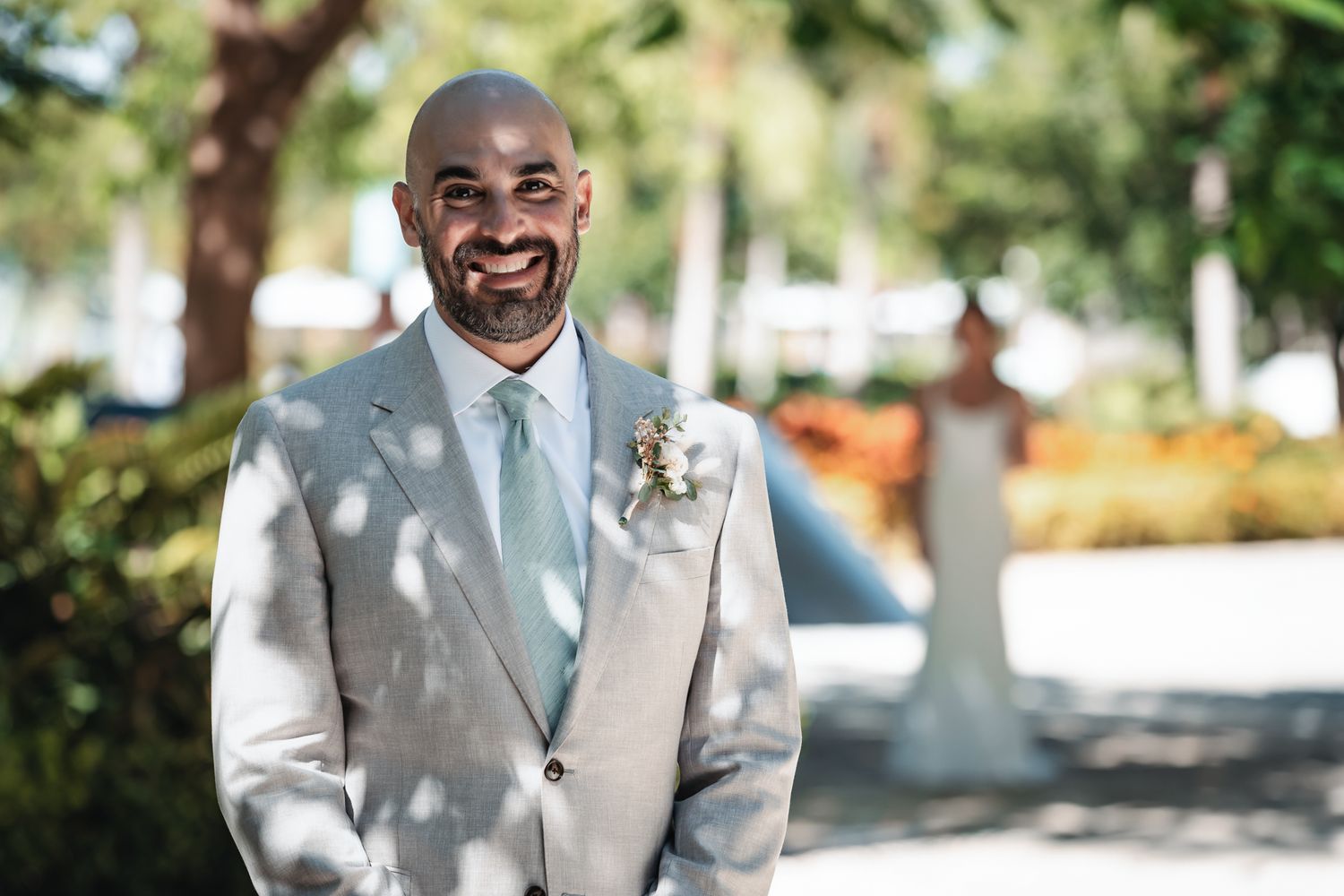 Smiling groom in light gray suit with mint tie and boutonniere, bride visible in background at outdoor wedding venue.