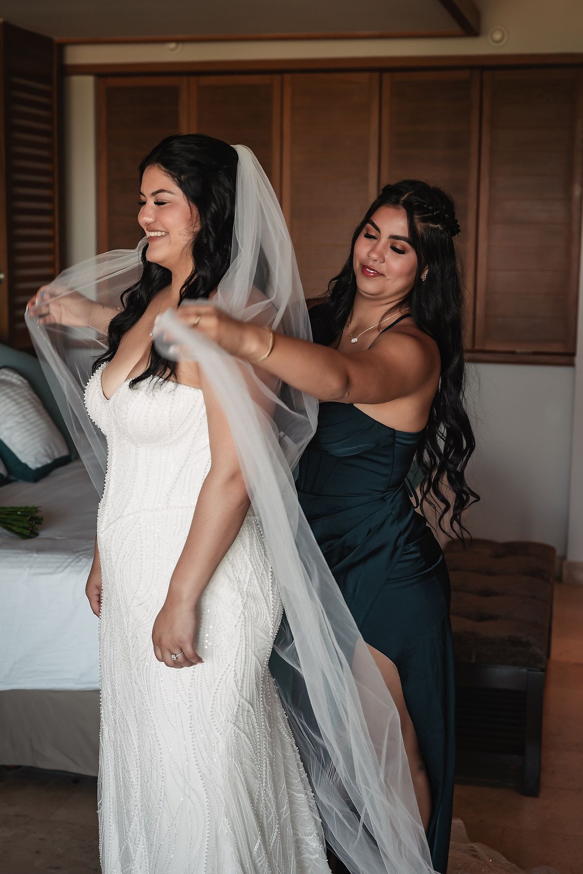 A bridesmaid in a dark green gown helps a smiling bride in a white lace gown arrange her flowing veil indoors.