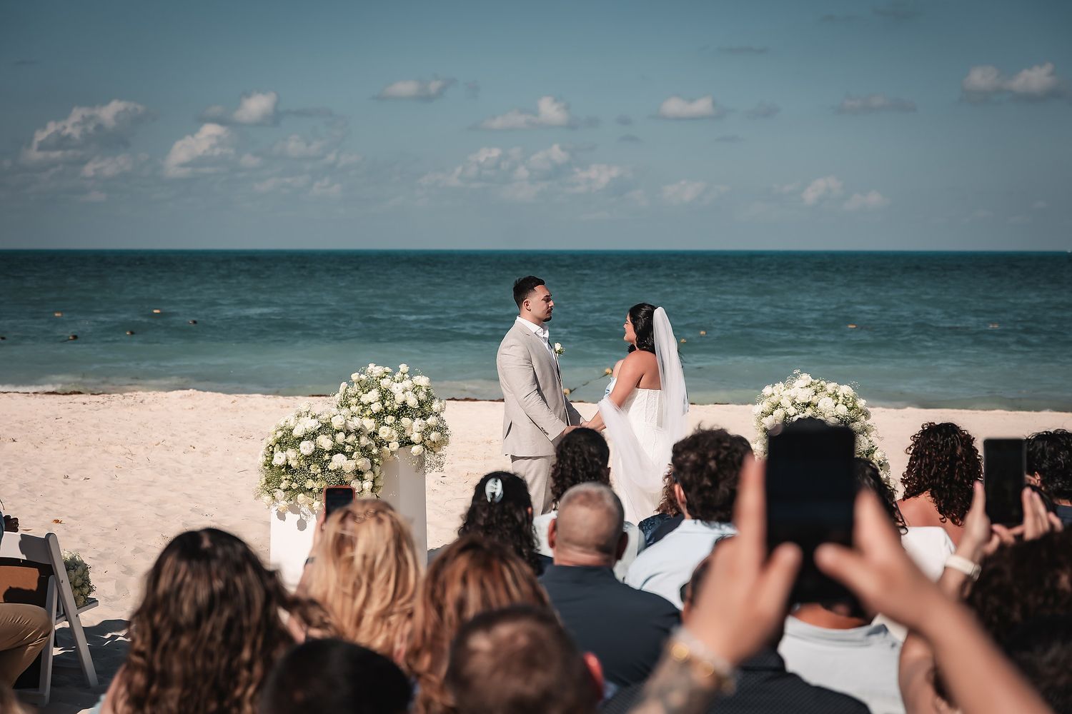 Couple exchanging vows at a romantic beach wedding ceremony with white floral arch and ocean backdrop.