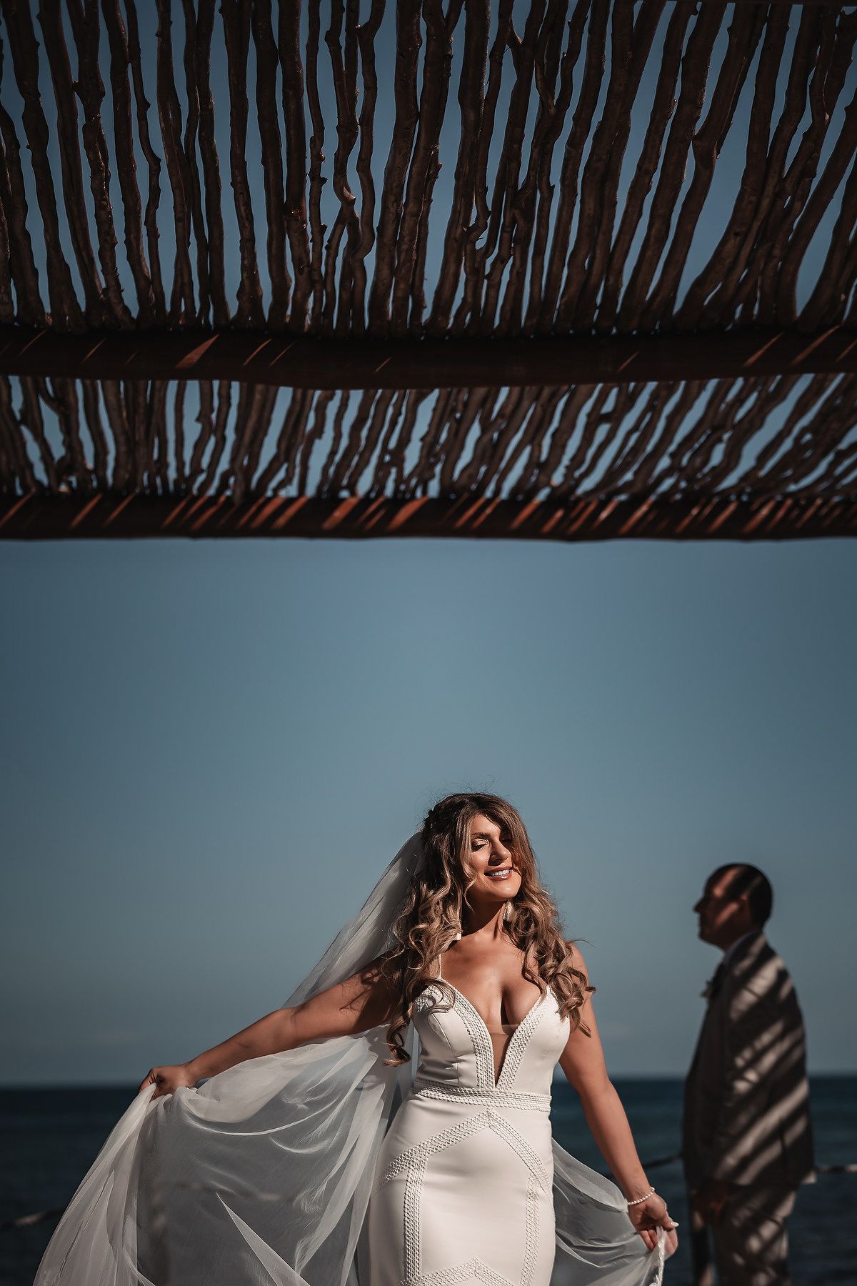 Bride in white gown and veil poses on beach under rustic wooden canopy, groom stands in background by ocean.