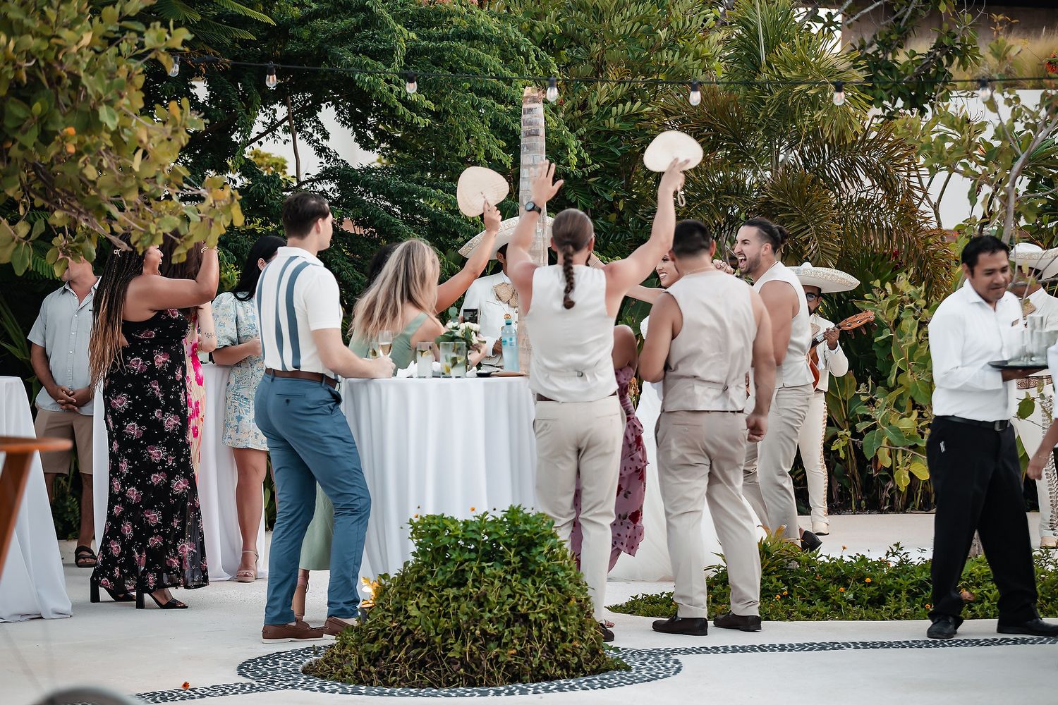 Guests celebrate outdoors at a tropical wedding reception, raising bouquets and dancing near a decorated white table.