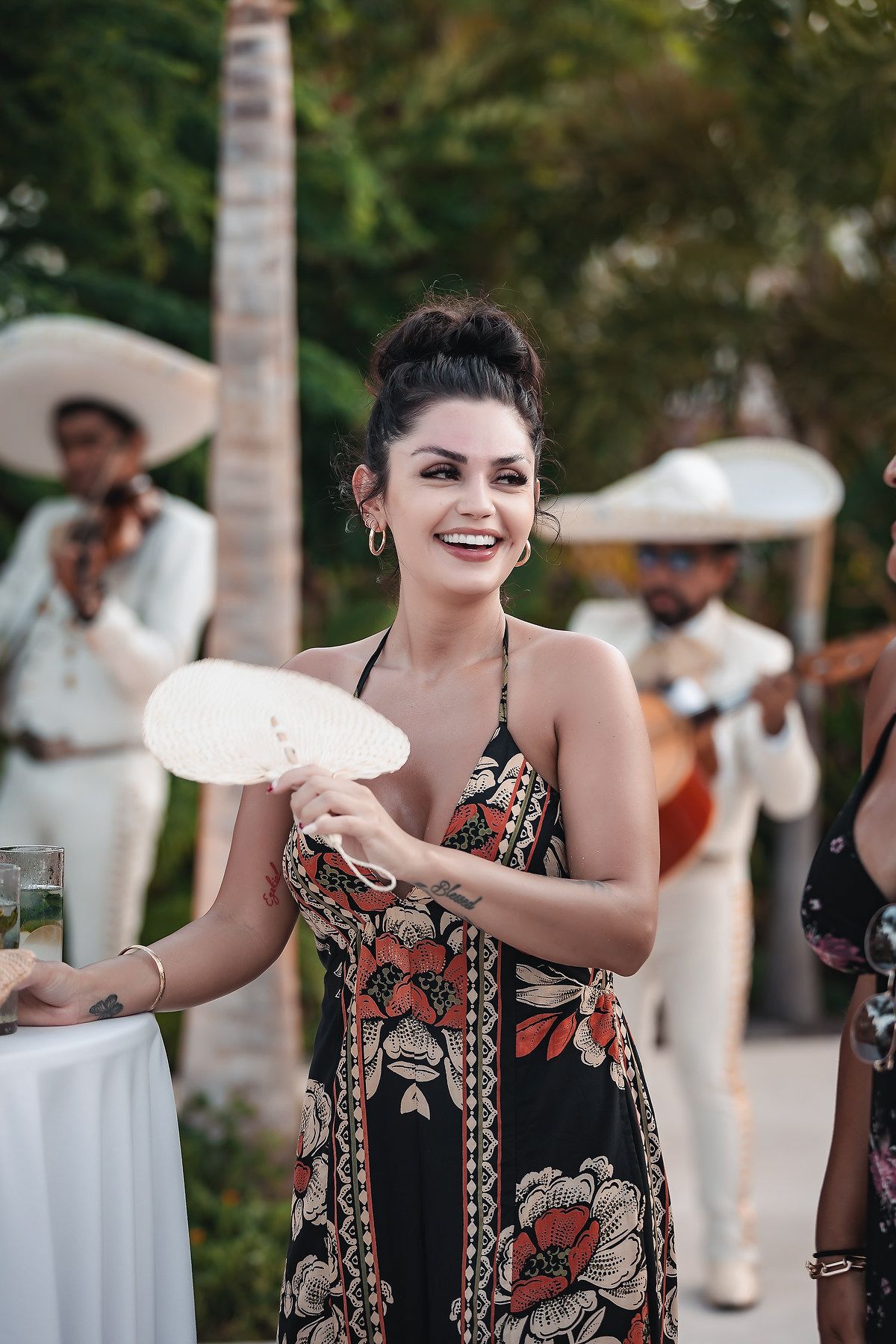 Woman in floral dress smiling and holding a fan at an outdoor Mexican mariachi party event.