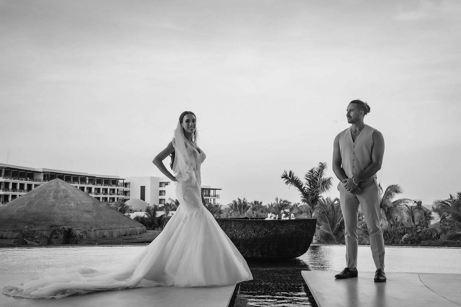 Black and white wedding photo of bride in mermaid gown and groom standing by a pool at a tropical resort.
