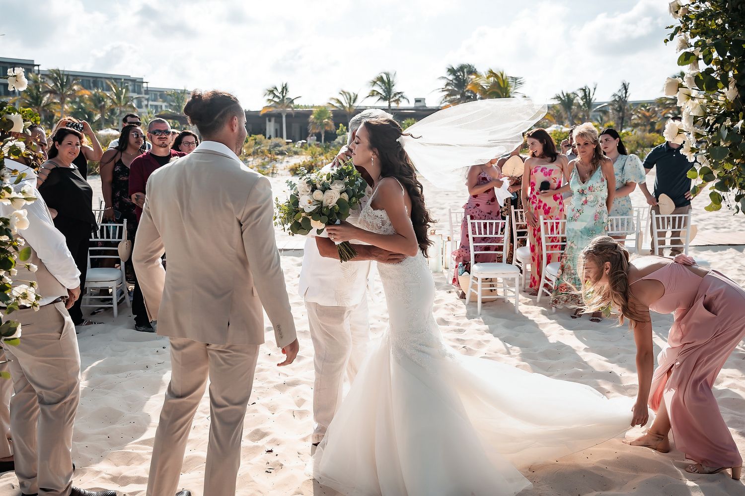 Bride walks down sandy beach aisle in white gown holding bouquet, groom waits as guests watch at outdoor wedding ceremony.