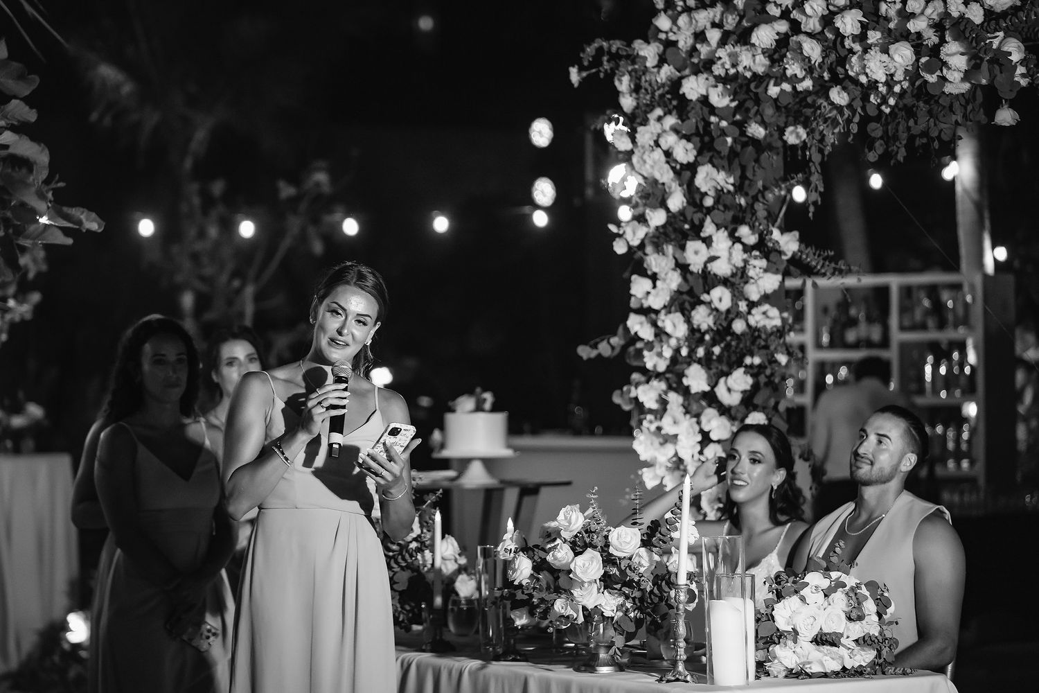 Bridesmaid delivers heartfelt speech at elegant outdoor wedding reception adorned with floral arrangements and string lights.