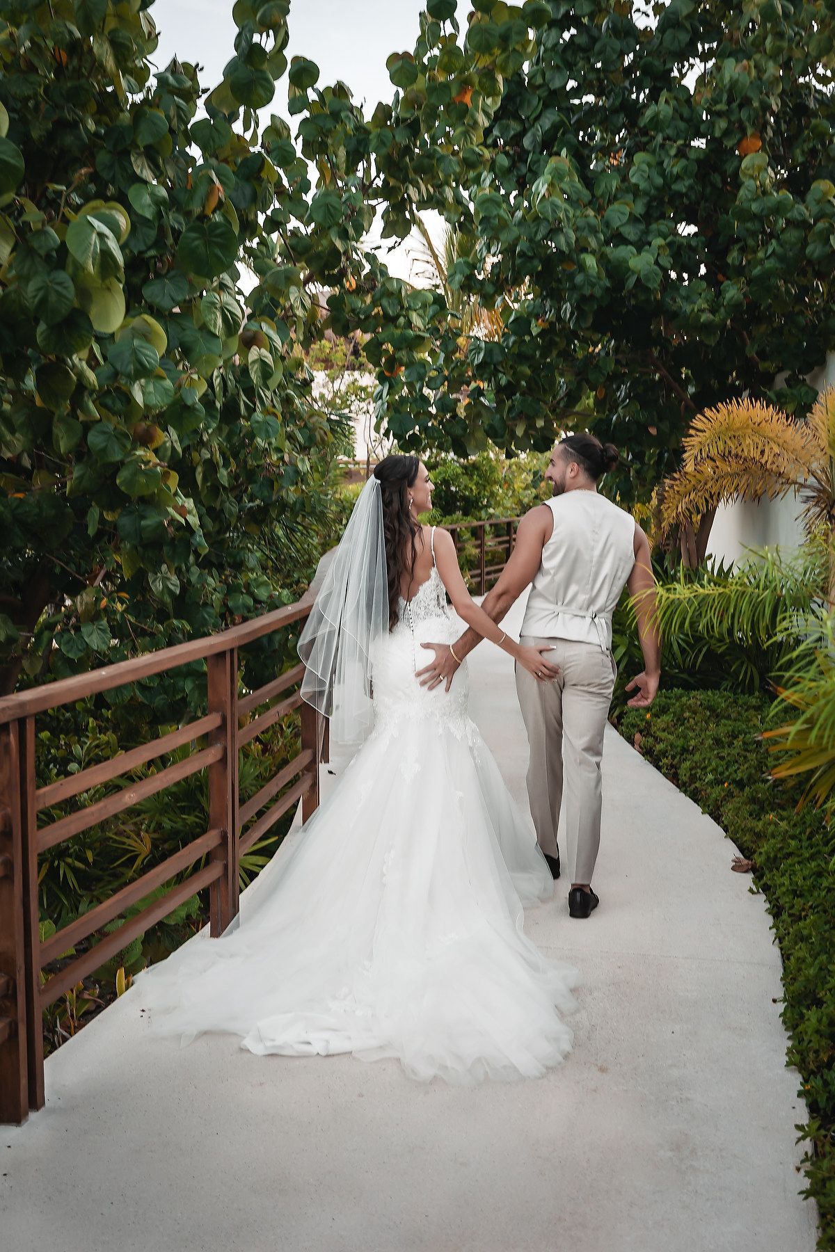 Bride and groom walking hand in hand down a tropical garden path, bride in white gown with veil.