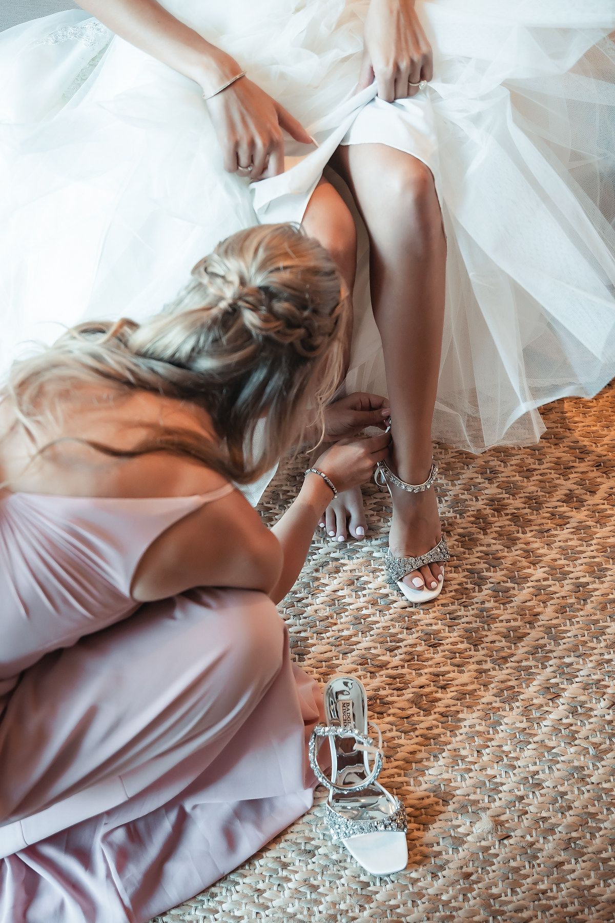 Bridesmaid in pink dress helping bride put on silver strappy heels before wedding ceremony.