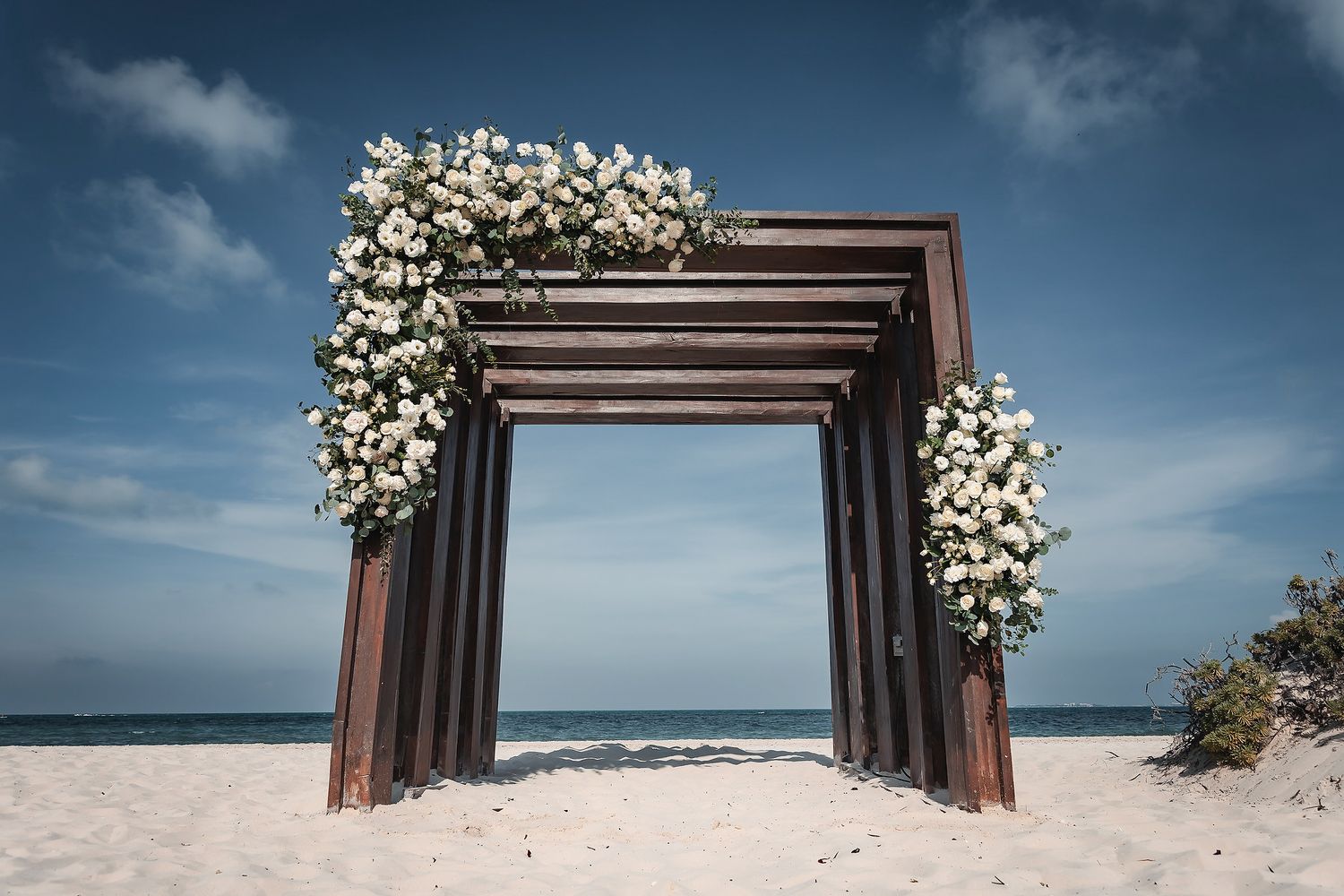 Elegant wooden beach wedding arch adorned with white floral arrangements on sandy shore under blue sky.