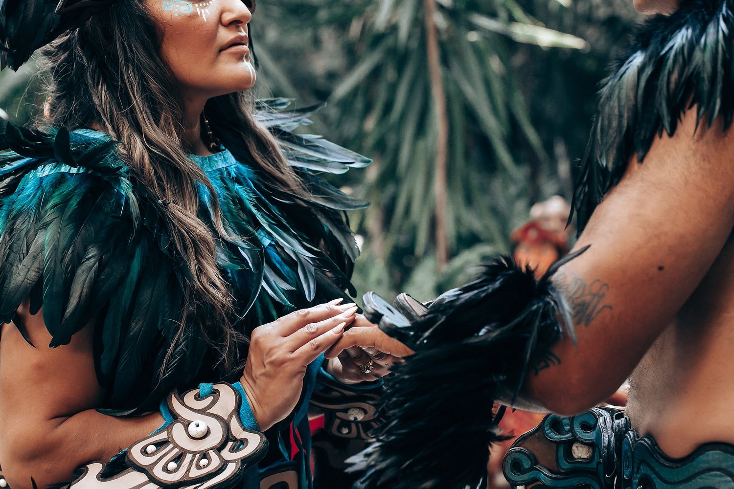 Woman in black feathered Mayan costume adjusts another performers outfit in a lush jungle setting.