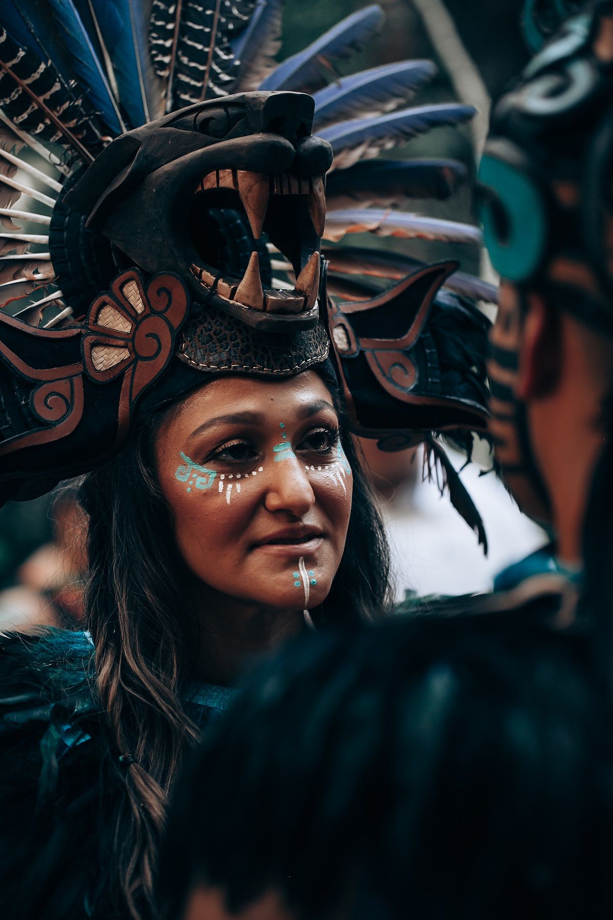 Close-up of a woman in an ornate Mayan headdress with turquoise face paint and decorative makeup.