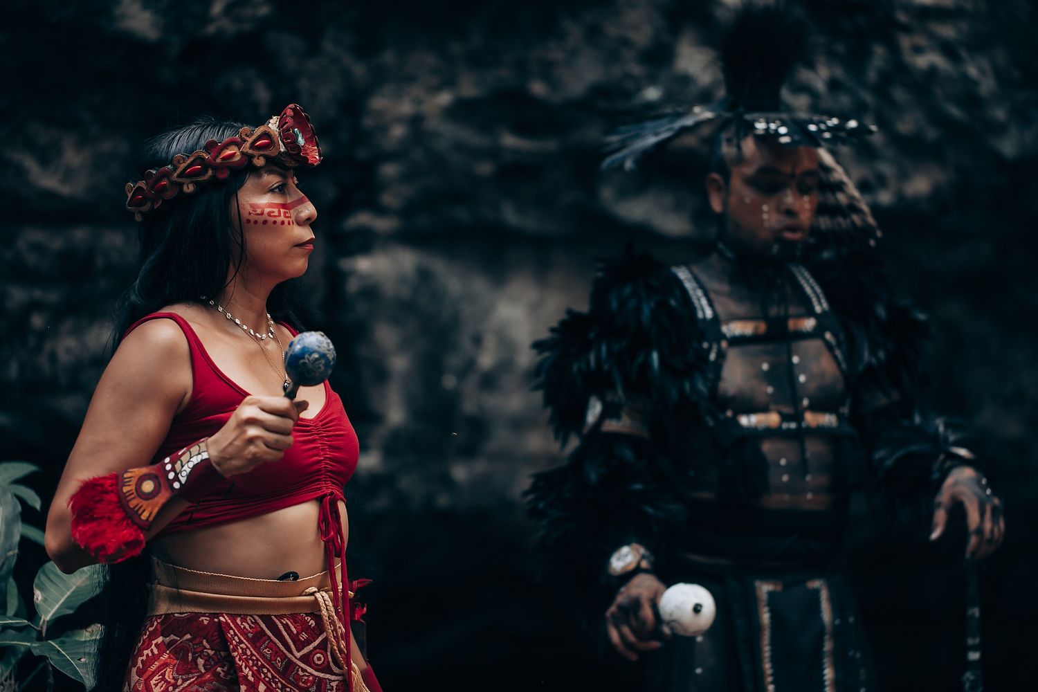 Two performers in traditional indigenous costumes with red attire and feathered armor pose dramatically outdoors.