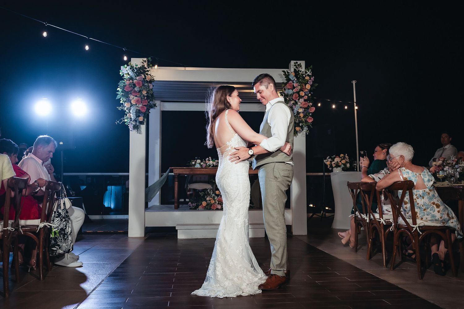 Newlyweds share their first dance outdoors at night under floral arch with string lights.