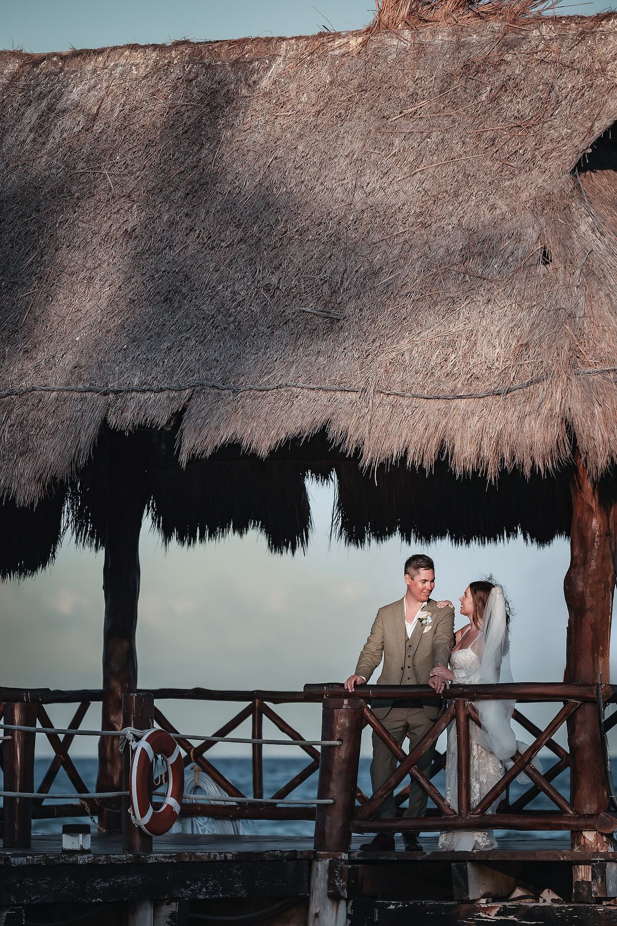 Bride and groom stand on a tropical pier gazebo with thatched roof overlooking the ocean at sunset.