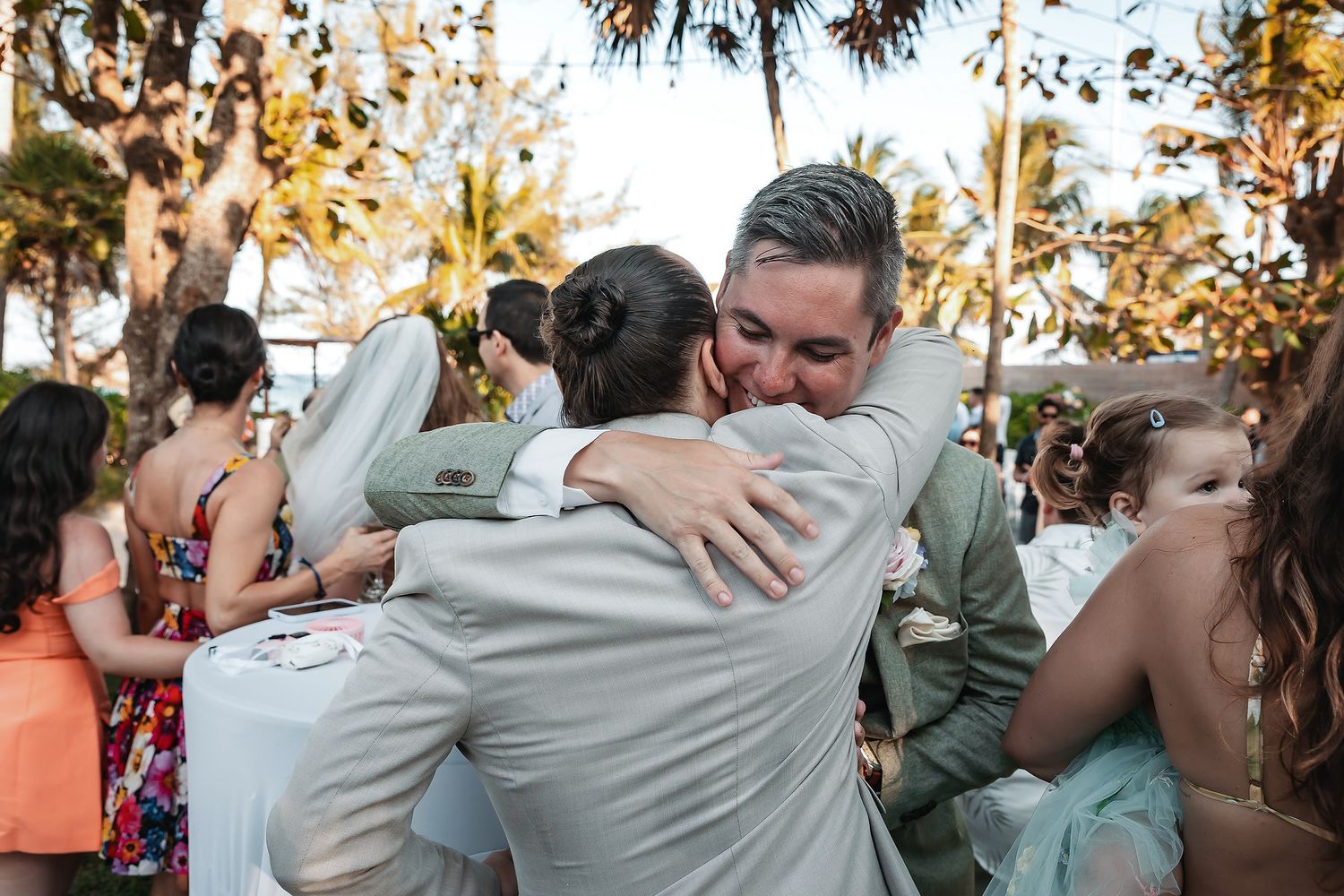 Two men share a heartfelt hug at an outdoor wedding reception surrounded by guests and palm trees.