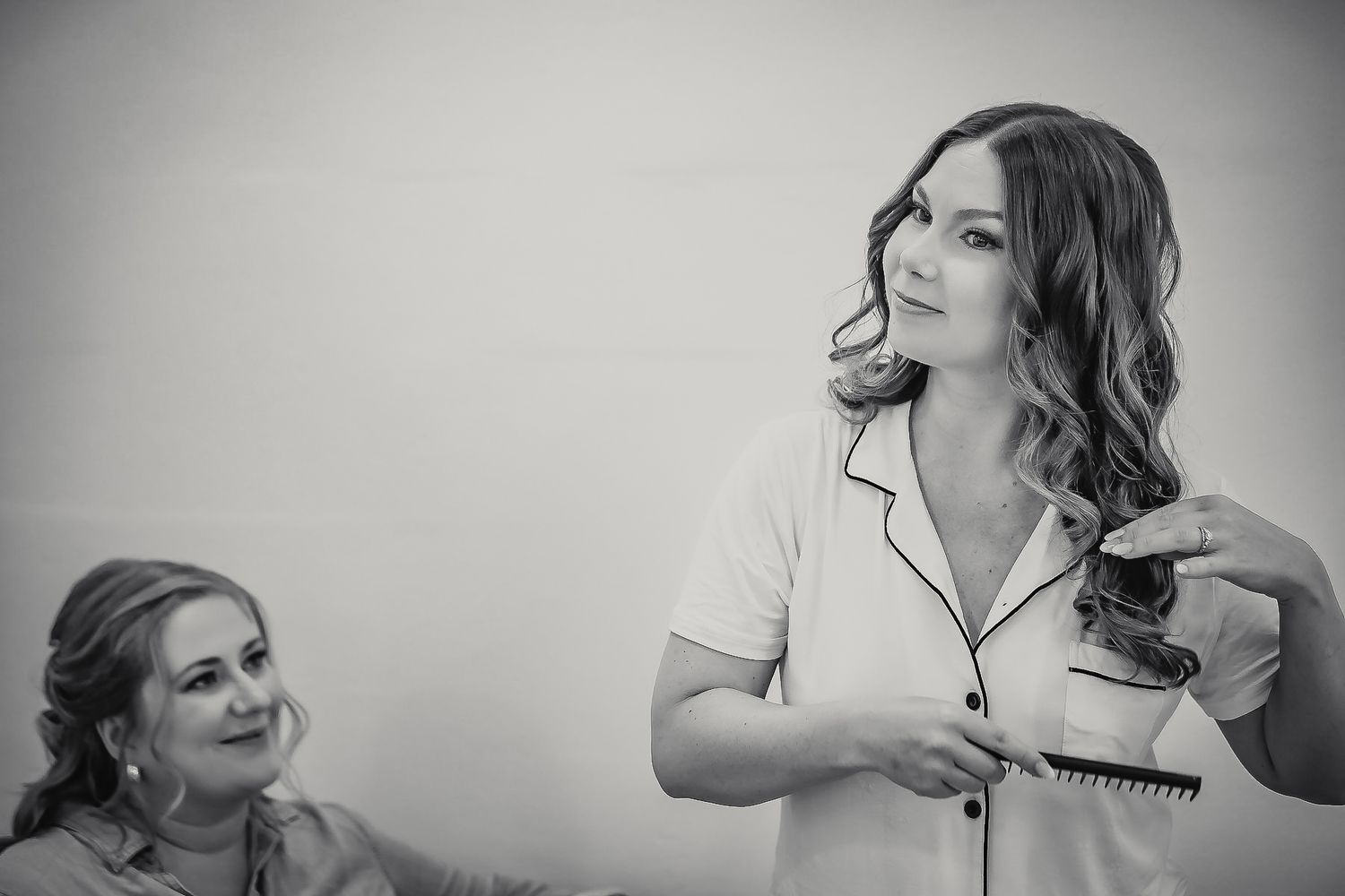 Black and white photo of a bride in pajamas curling her hair while a bridesmaid watches and smiles during wedding preparations.