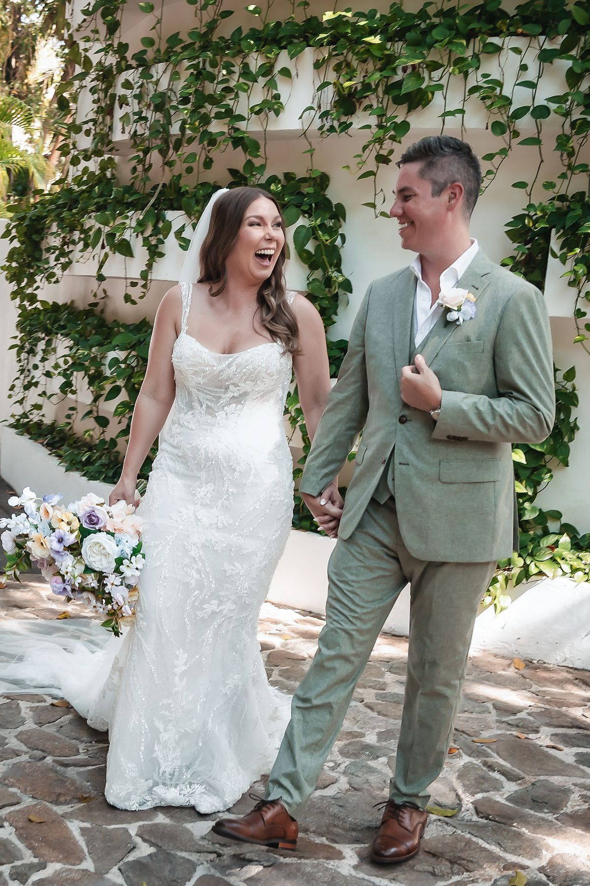 Joyful bride in white lace gown and groom in sage green suit hold hands, laughing, in front of ivy-covered wall.