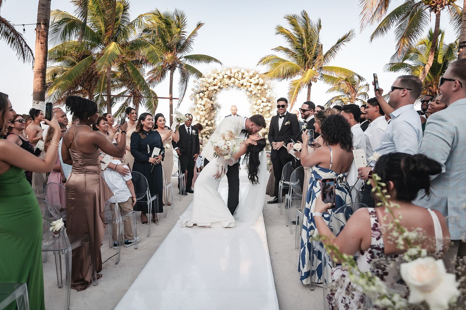 Bride and groom kiss at outdoor beach wedding ceremony with floral arch, palm trees, and cheering guests lining white aisle.