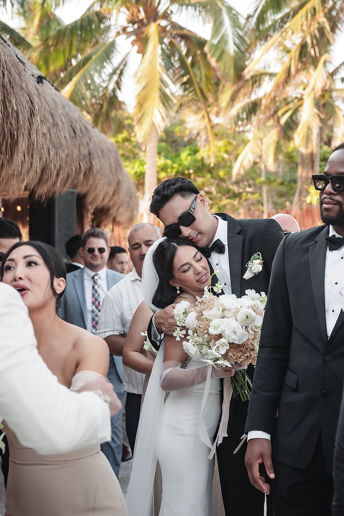 Bride and groom share a tender embrace at tropical beach wedding, surrounded by guests with palm trees in background.