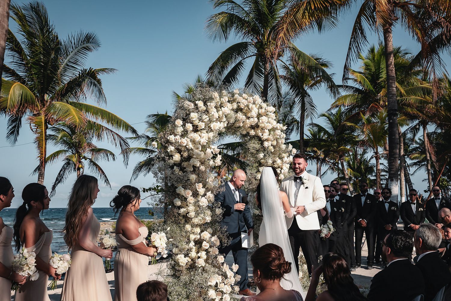 Beach wedding ceremony with floral arch, groom awaiting bride, bridesmaids, palm trees, and ocean backdrop.