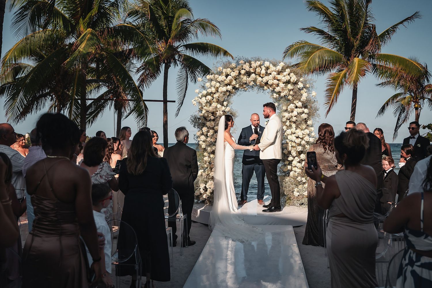 Outdoor beach wedding ceremony under floral arch with palm trees, couple exchanging vows before guests.