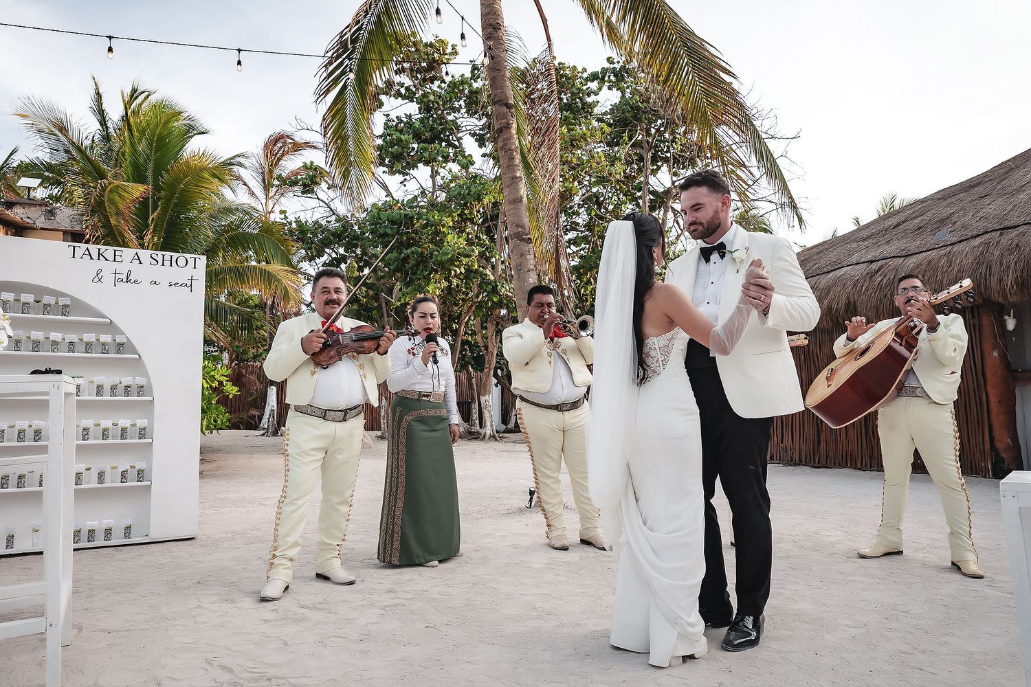 Bride and groom dance on beach while mariachi band plays at tropical outdoor wedding celebration.