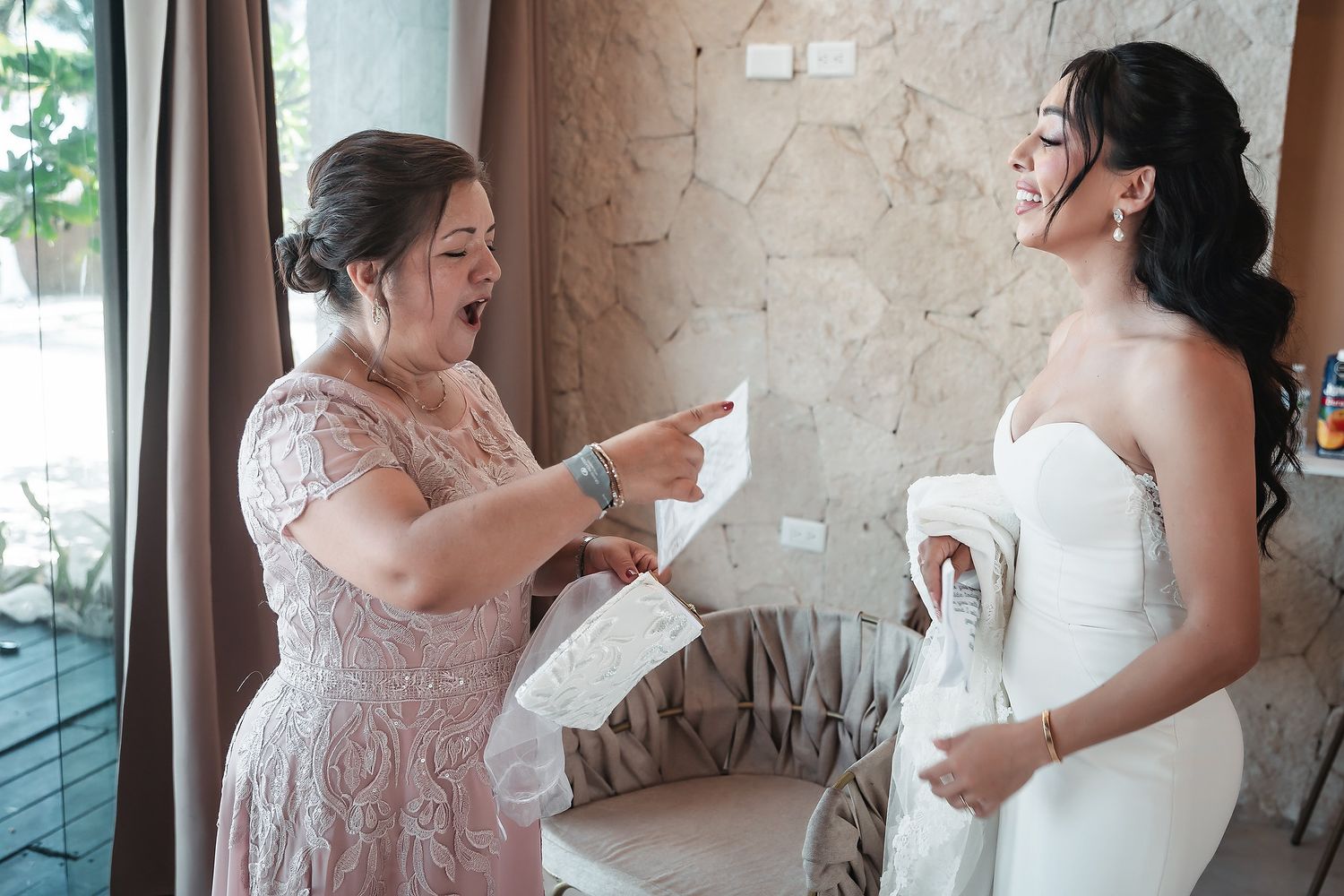 Bride in white strapless gown laughs with woman in pink lace dress during wedding preparations indoors.