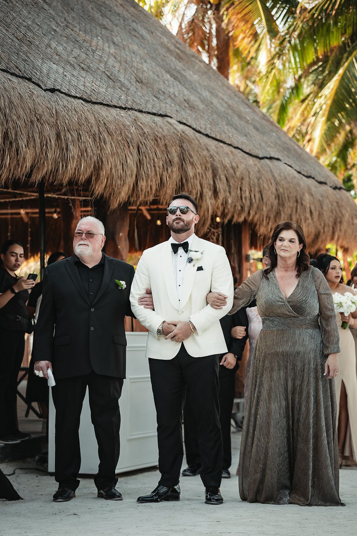 Groom in white tuxedo jacket walks down aisle with parents at tropical beach wedding ceremony.