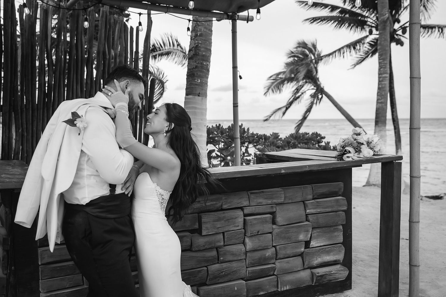 Bride and groom share a romantic kiss at a beachside stone bar with palm trees in black and white.