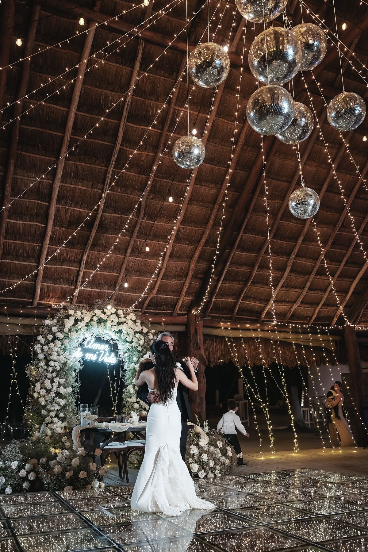 Bride and groom dance under sparkling disco balls and string lights in a rustic wedding venue with floral arch.